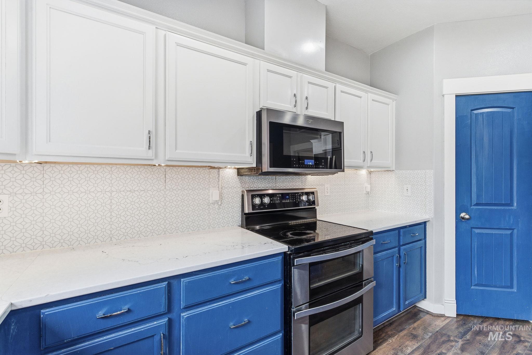 Kitchen with blue cabinets, stainless steel appliances, white cabinets, dark wood-style floors, and decorative backsplash